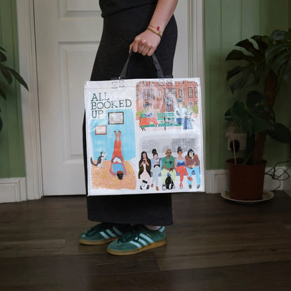 A person standing indoors holding a tote bag printed with an illustrated cover design that reads 'ALL BOOKED UP.' The scene includes a green wall, white door, wood floor, and potted plant in the background.