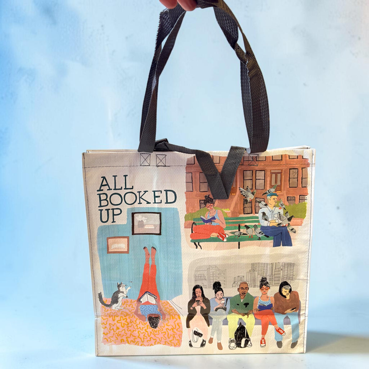 A canvas tote bag with black handles, featuring a collage of illustrated scenes and the text 'ALL BOOKED UP' on a light blue background.