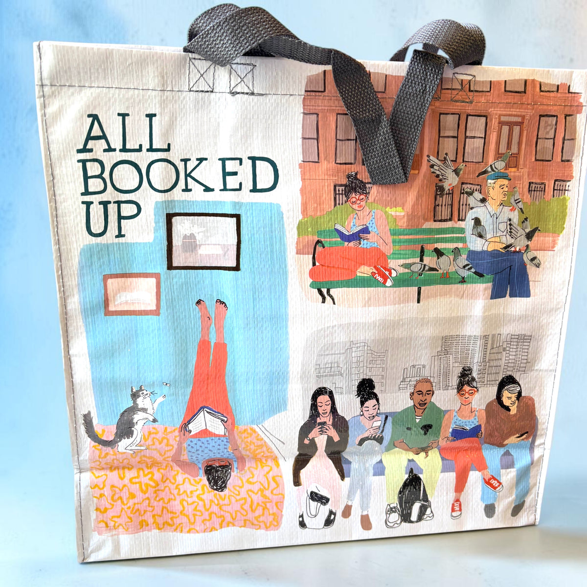 A canvas tote bag with dark gray webbed handles, featuring a collage-style illustration with the text 'ALL BOOKED UP' and scenes of people, pets, and buildings. The bag is photographed against a light blue background.