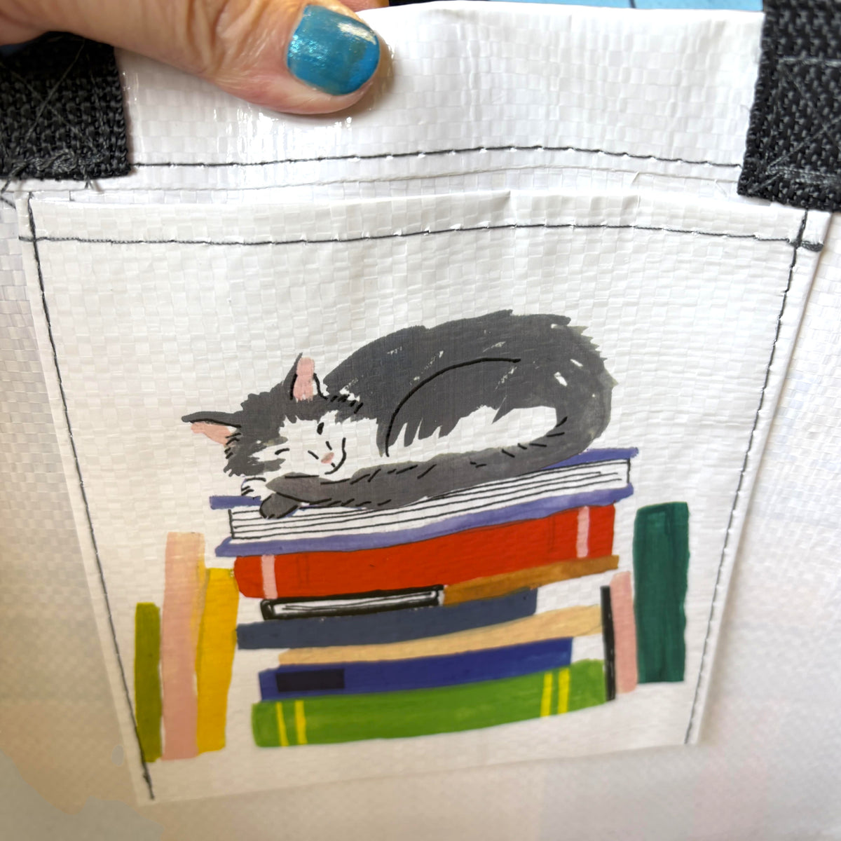 A close-up of a white canvas tote bag with a front pocket featuring a painted illustration of a sleeping gray and white cat resting on a stack of colorful books. A hand with blue glitter nail polish is holding the top edge of the bag.