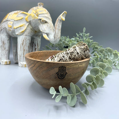 Wooden bowl with sage and a Hamsa hand symbol, next to a decorative elephant and eucalyptus leaves on a light background
