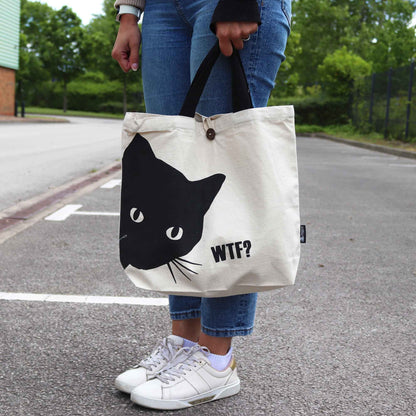 Person holding a tote bag with a black cat design and 'WTF?' text on a street.