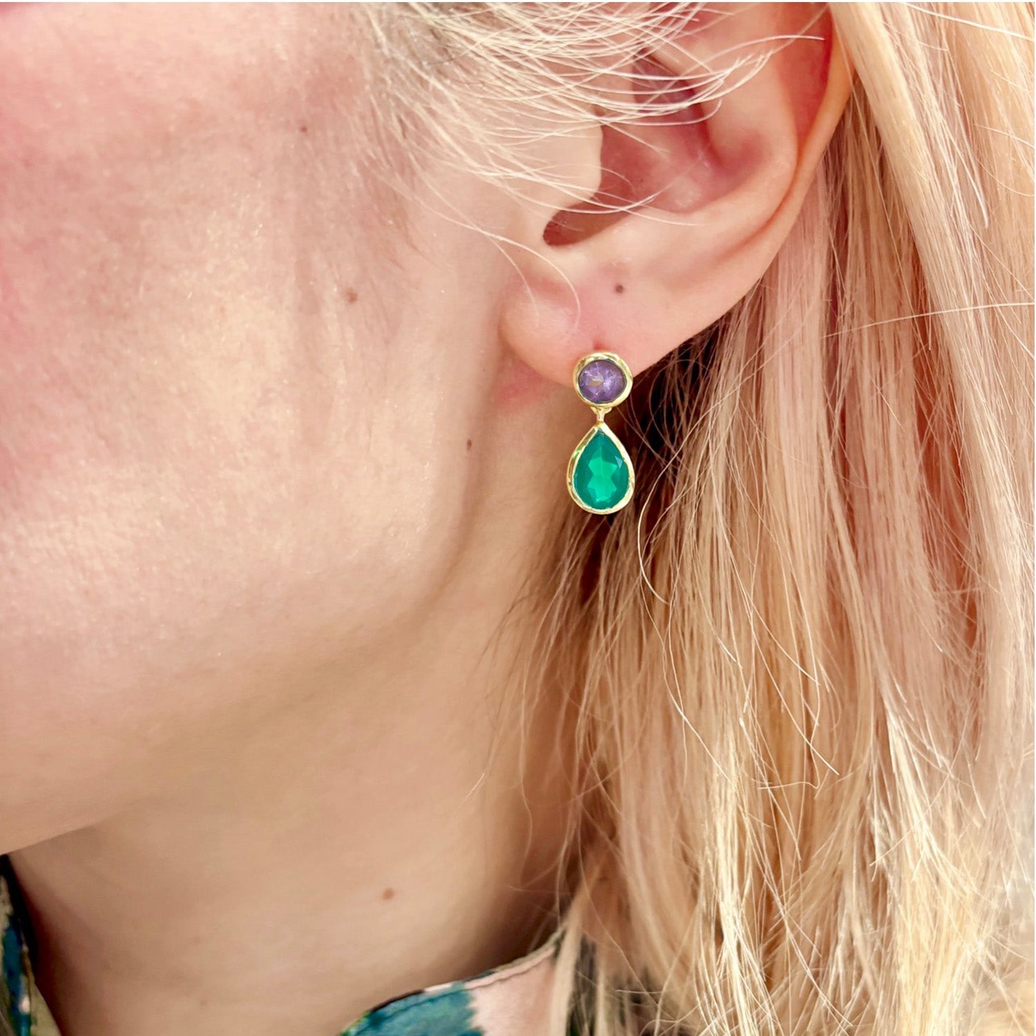 Close-up of an ear wearing a gold earring with green and purple gemstones.