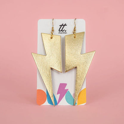 Gold lightning bolt earrings on a colorful card with 'tt.' brand name against a pink background