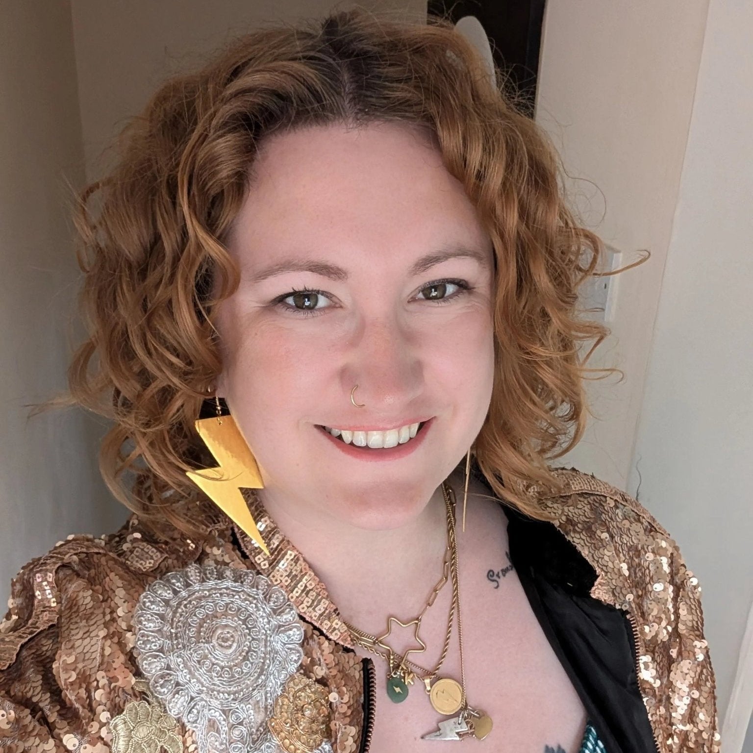 Woman with curly hair wearing a gold sequin jacket and multiple necklaces.