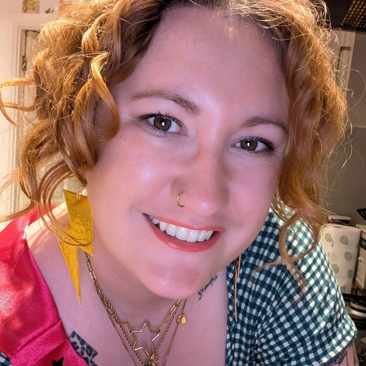 Woman with curly hair wearing a green checkered shirt and yellow earrings indoors.