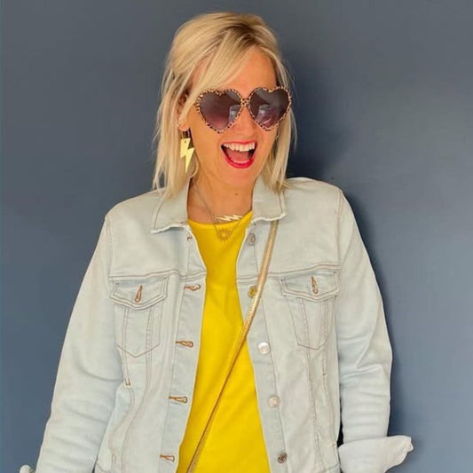 Woman wearing a light blue denim jacket and yellow top with heart-shaped sunglasses against a gray background