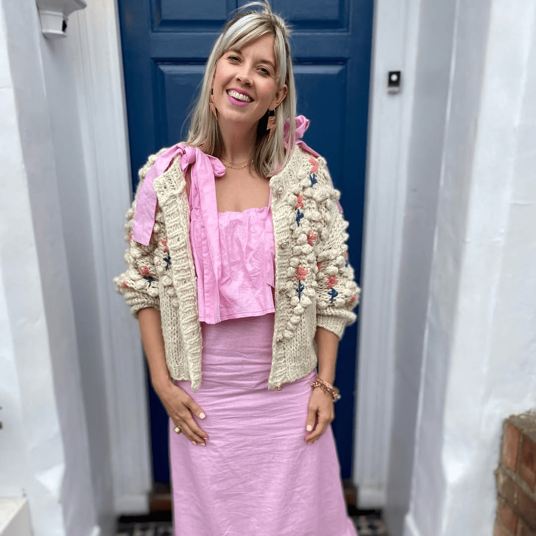 Woman wearing a pink top and skirt with a beige cardigan in front of a blue door.