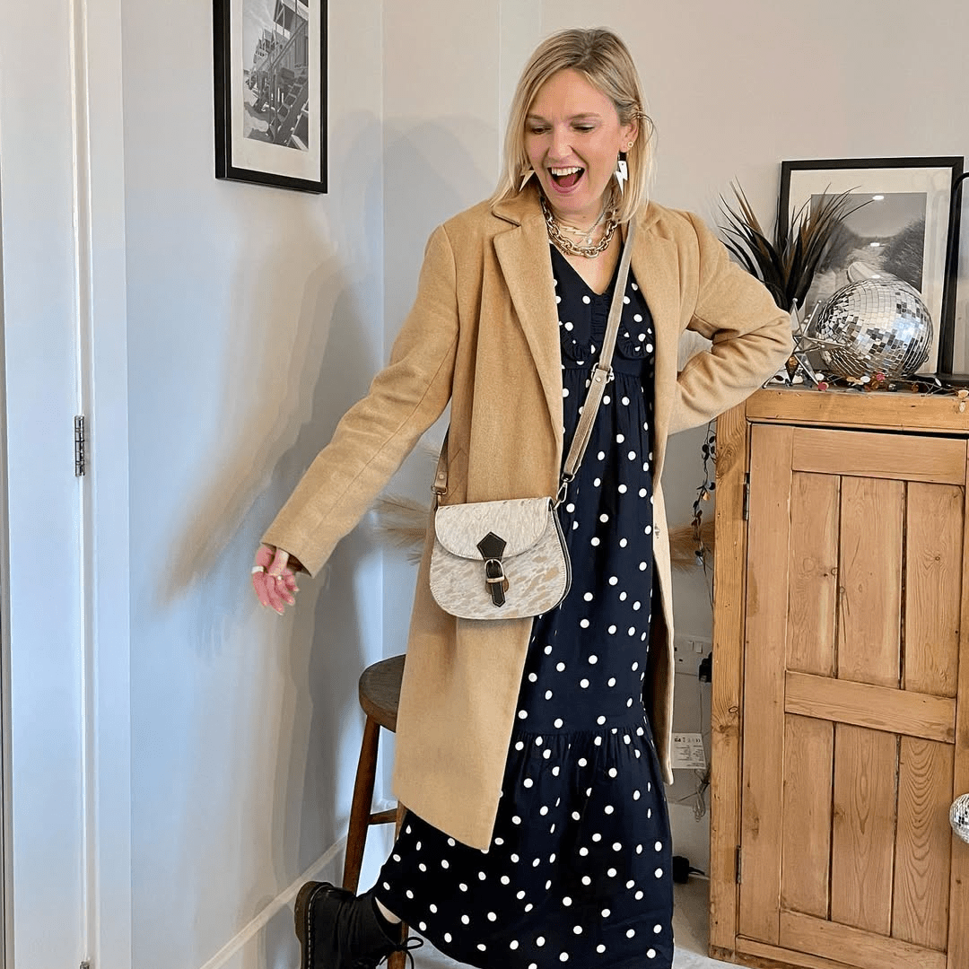 Woman in a beige coat and polka dot dress standing in a room with wooden furniture and decor.
