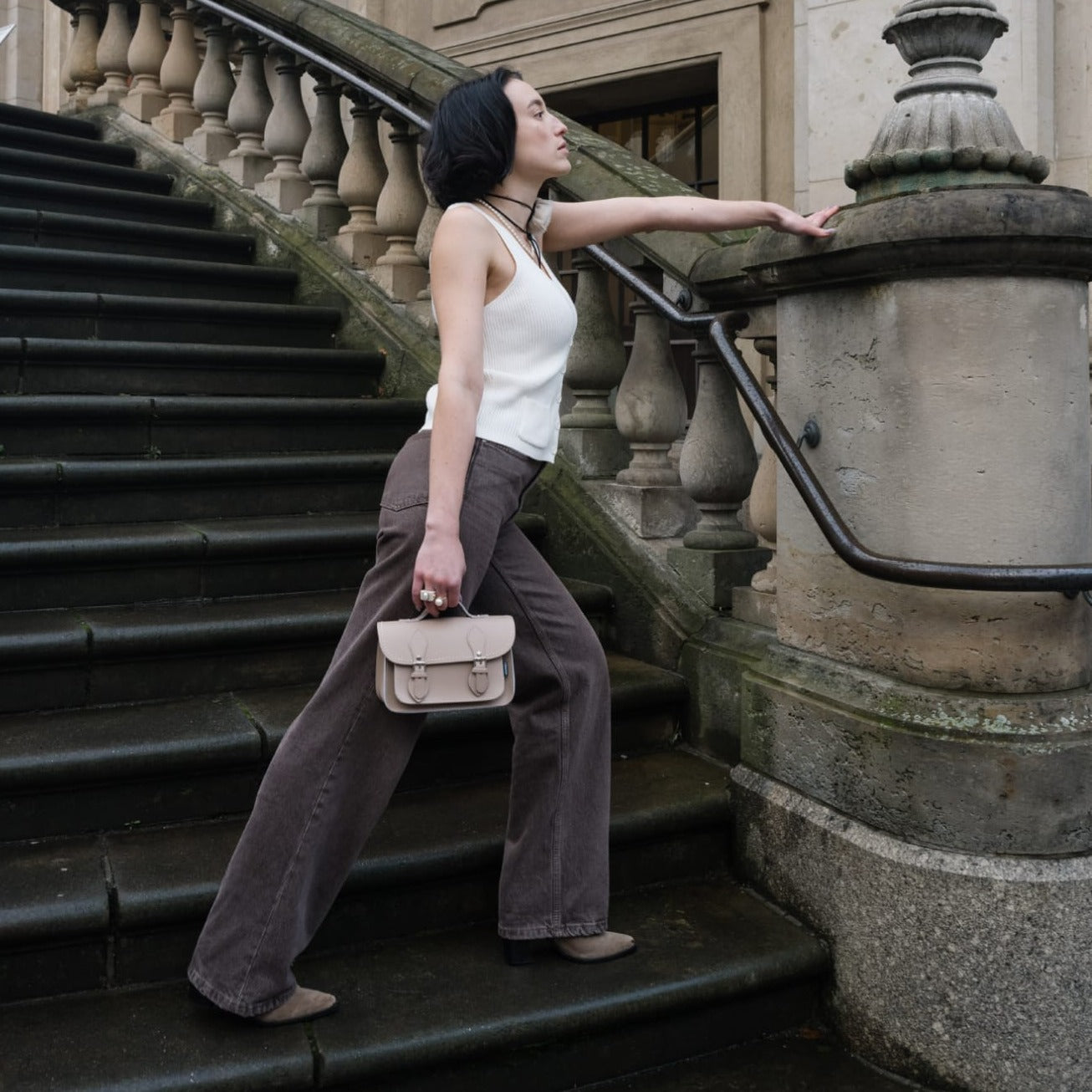 Woman standing on a set of stairs holding a handbag, wearing a white top and gray pants.