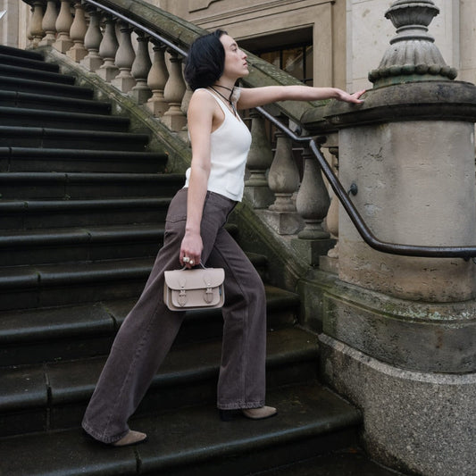 Woman standing on a set of stairs holding a handbag, wearing a white top and gray pants.