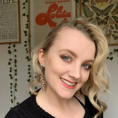 Woman with blonde hair and blue eyes smiling in front of a 'Lolita' poster and butterfly artwork.