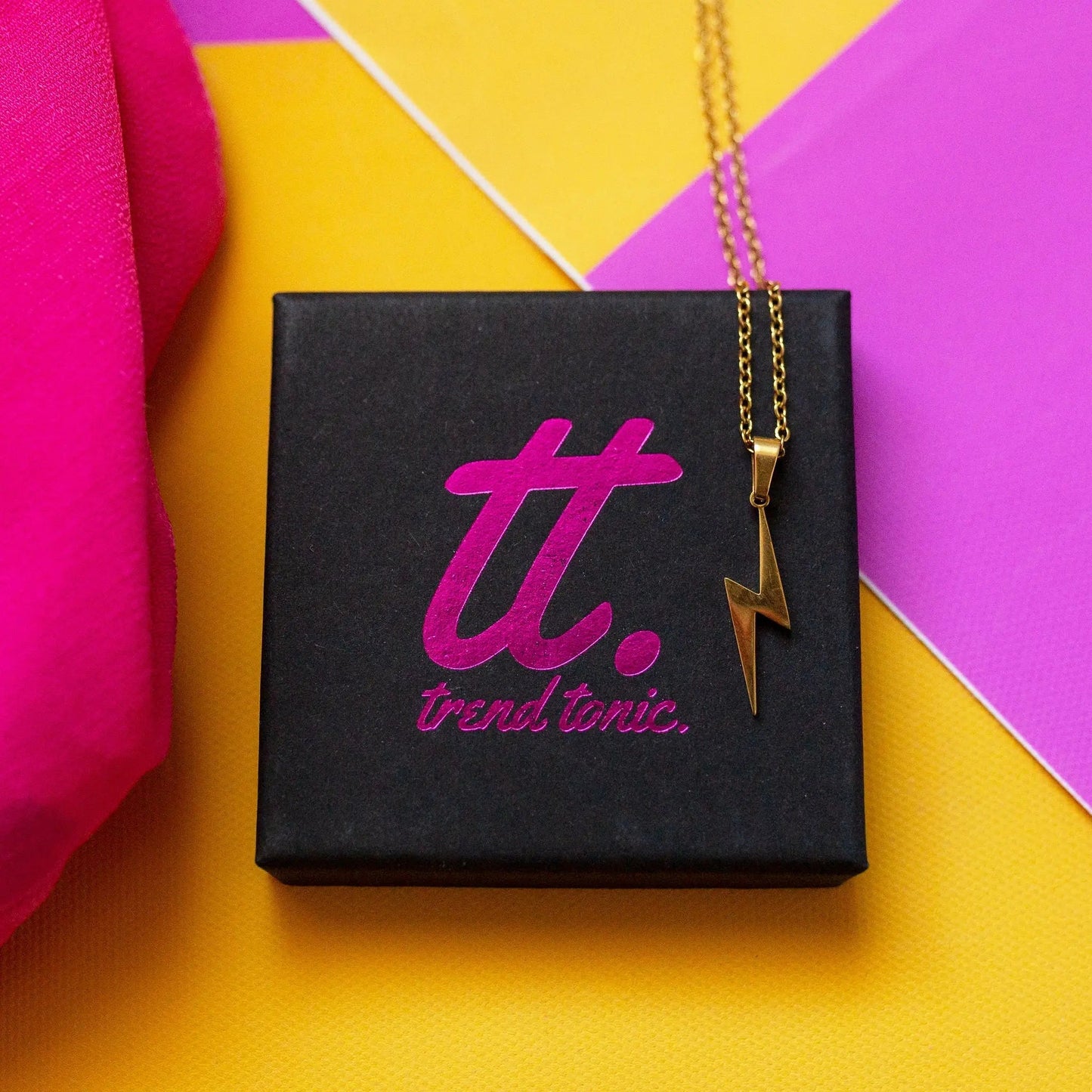 Gold necklace with a lightning bolt pendant on a Trendylicious box against a colorful background