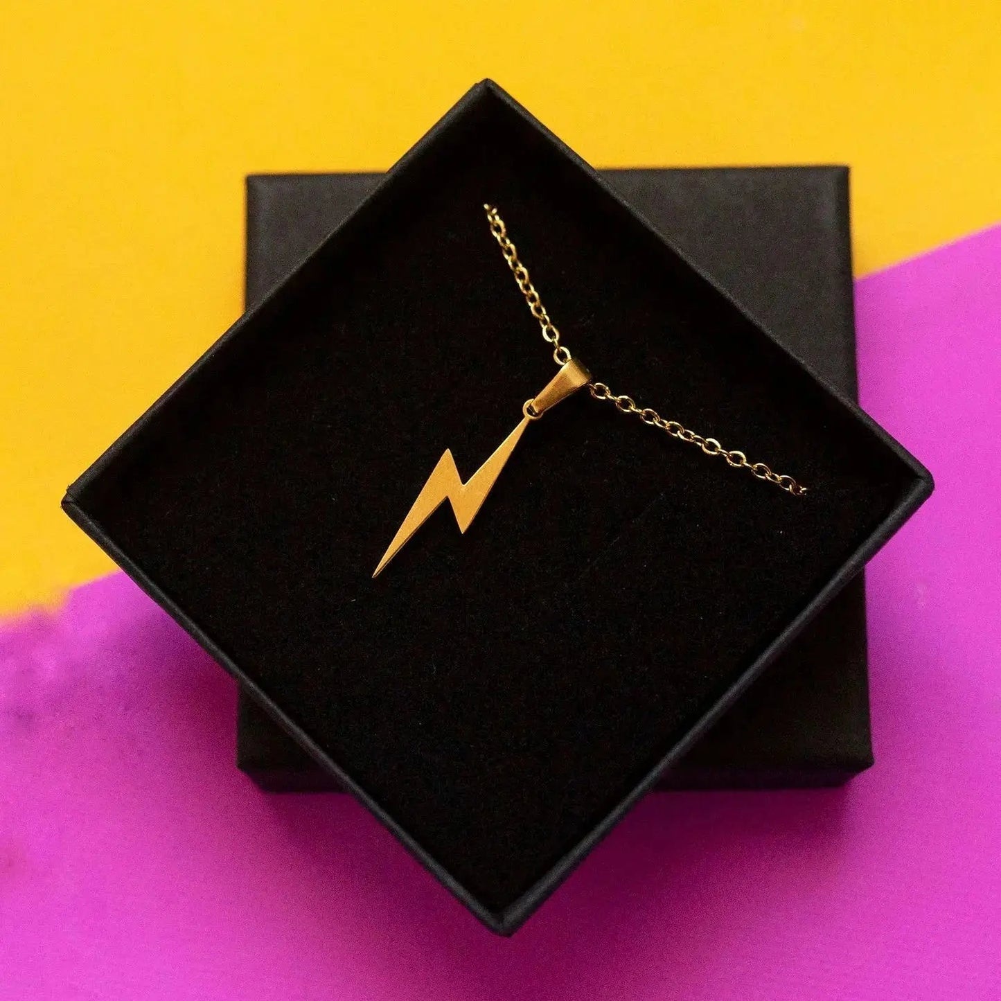 Gold necklace with a lightning bolt pendant on a black box against a yellow and purple background