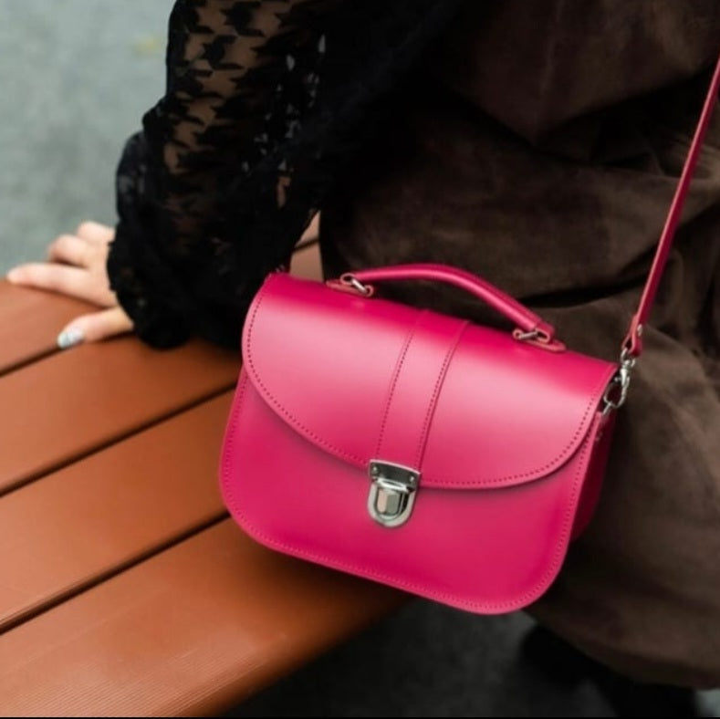 Pink handbag on a wooden surface with a person wearing a brown coat and black lace sleeves.