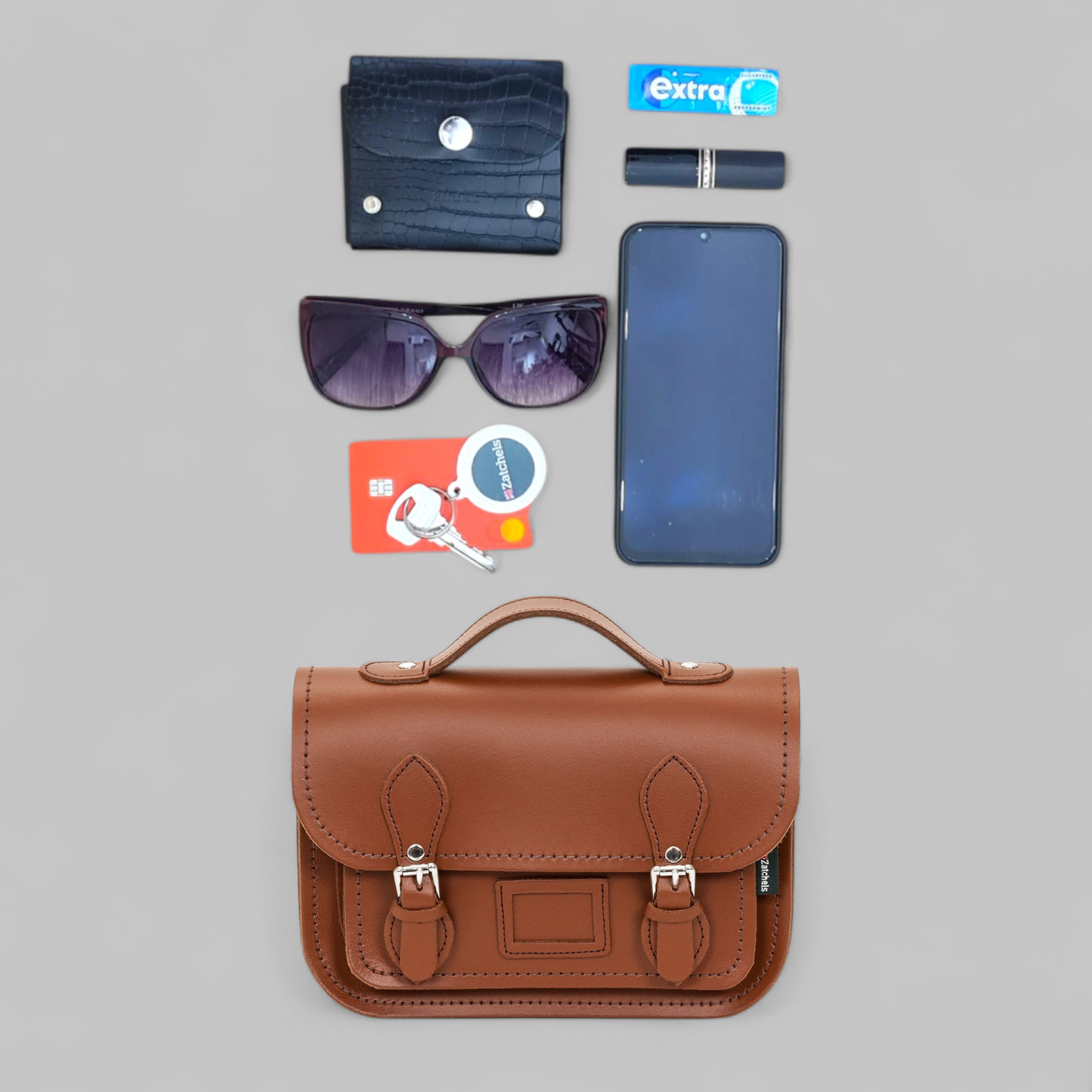 Collection of items including a brown leather satchel, sunglasses, phone, card, and wallet on a gray background.