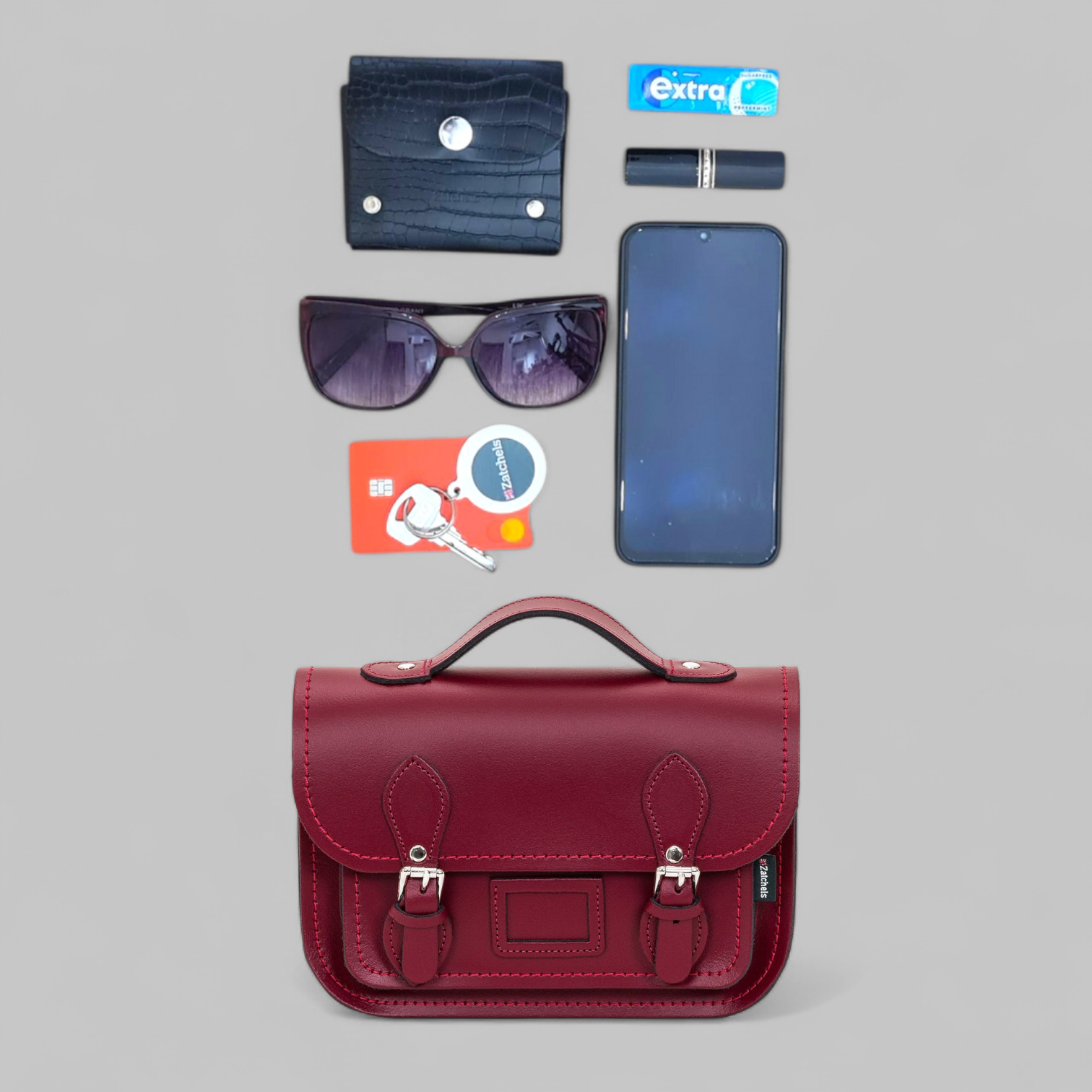 Red satchel bag with phone, sunglasses, wallet, and card on a gray background