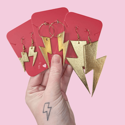 Gold lightning bolt earrings on red cards held by a hand with a pink background