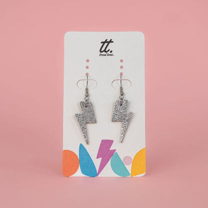 Pair of silver glittery lightning bolt earrings on a branded card against a pink background