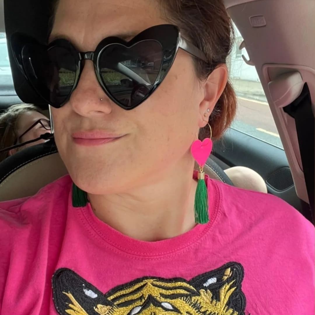 Person wearing heart-shaped sunglasses and a pink shirt with a tiger design inside a vehicle.