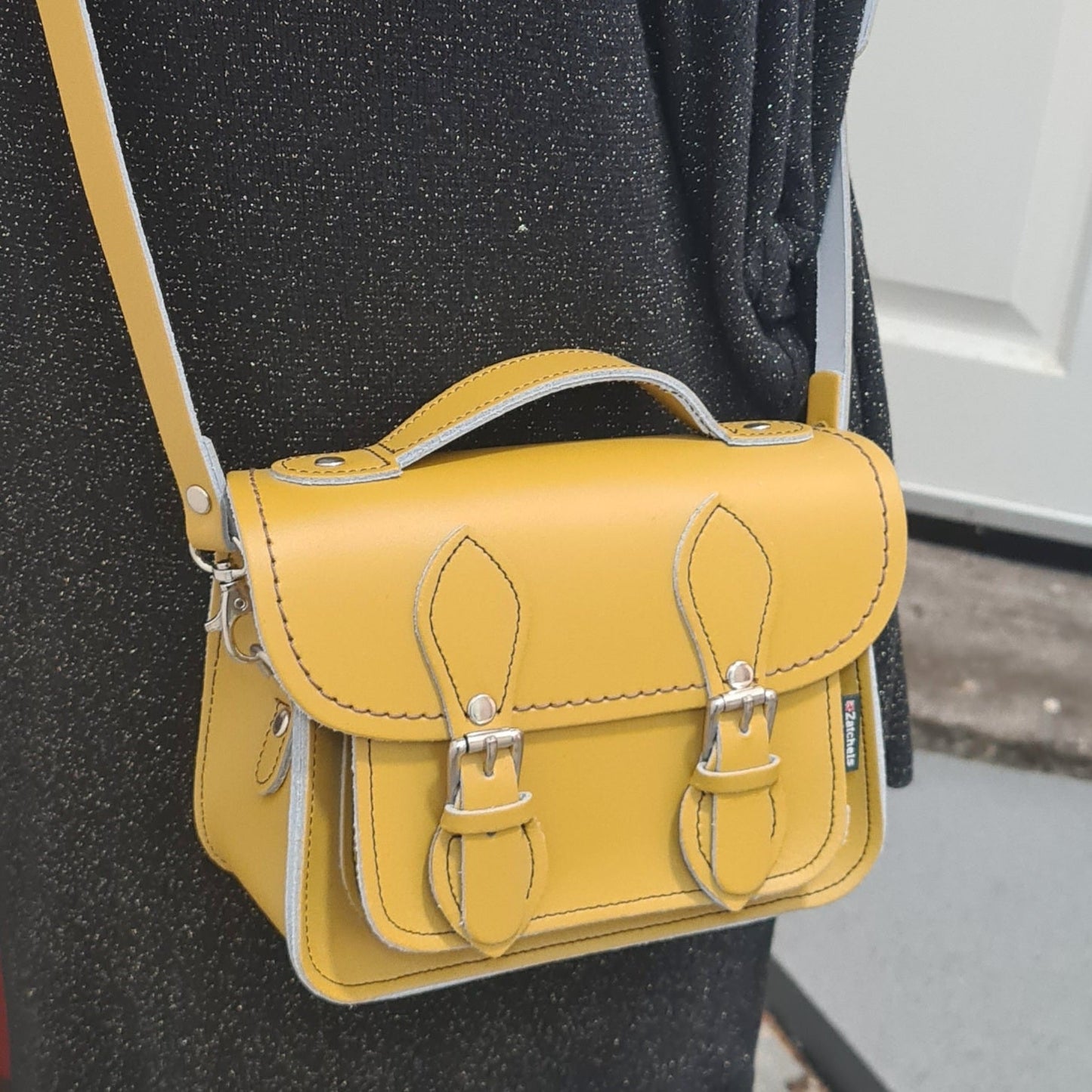 Yellow satchel bag with silver buckles worn by a person against a neutral background