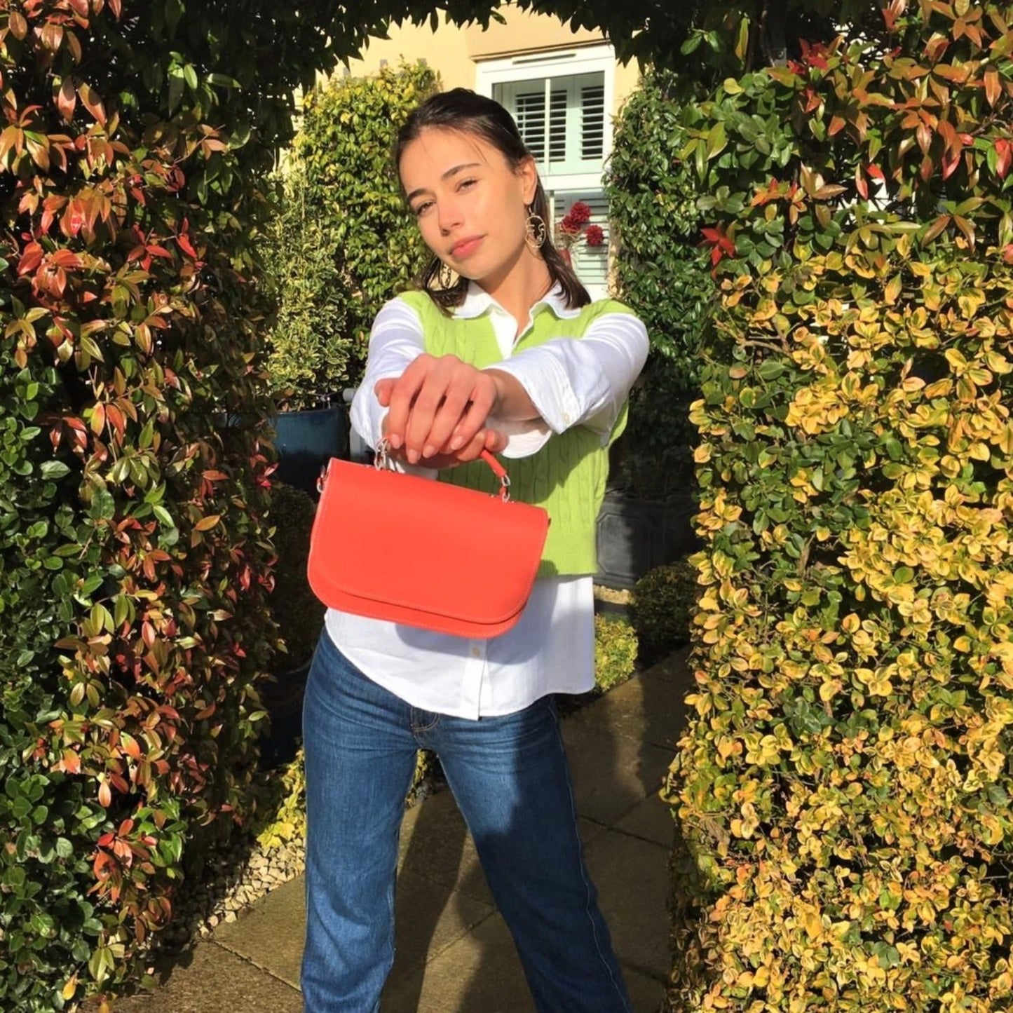 Woman holding a red handbag in front of green and yellow leafy bushes