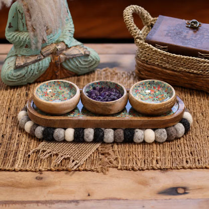 Grey felt ball oval mat under a wooden tray holding three small bowls, on a woven natural mat