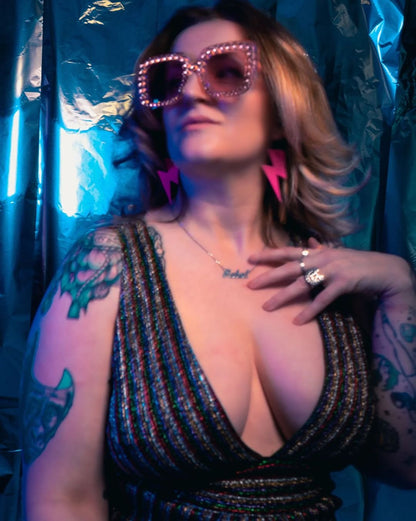 Person wearing sparkly sunglasses and a black dress with a deep neckline, standing against a blue curtain.