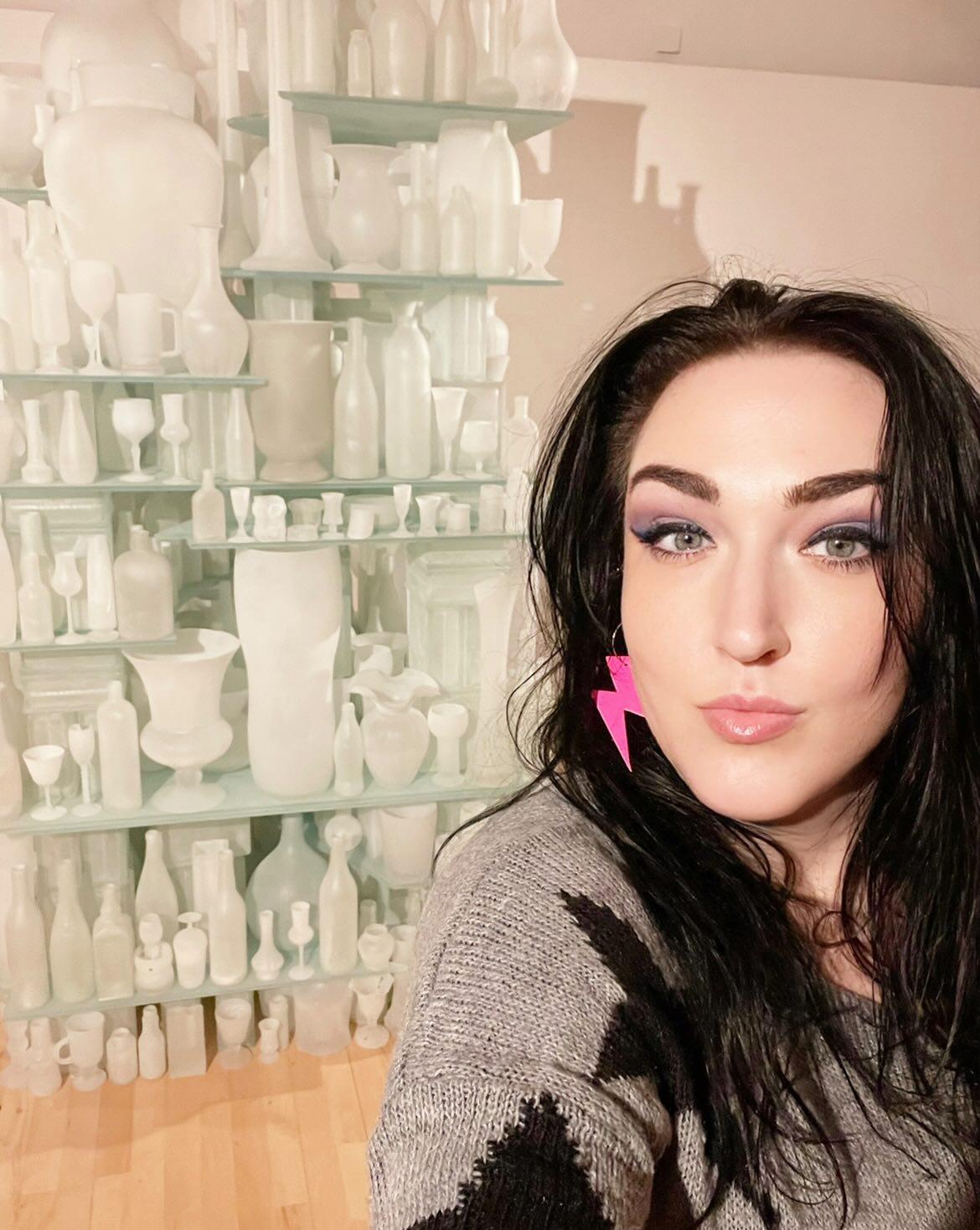 Woman with dark hair and pink earrings in front of a shelf with white ceramic items