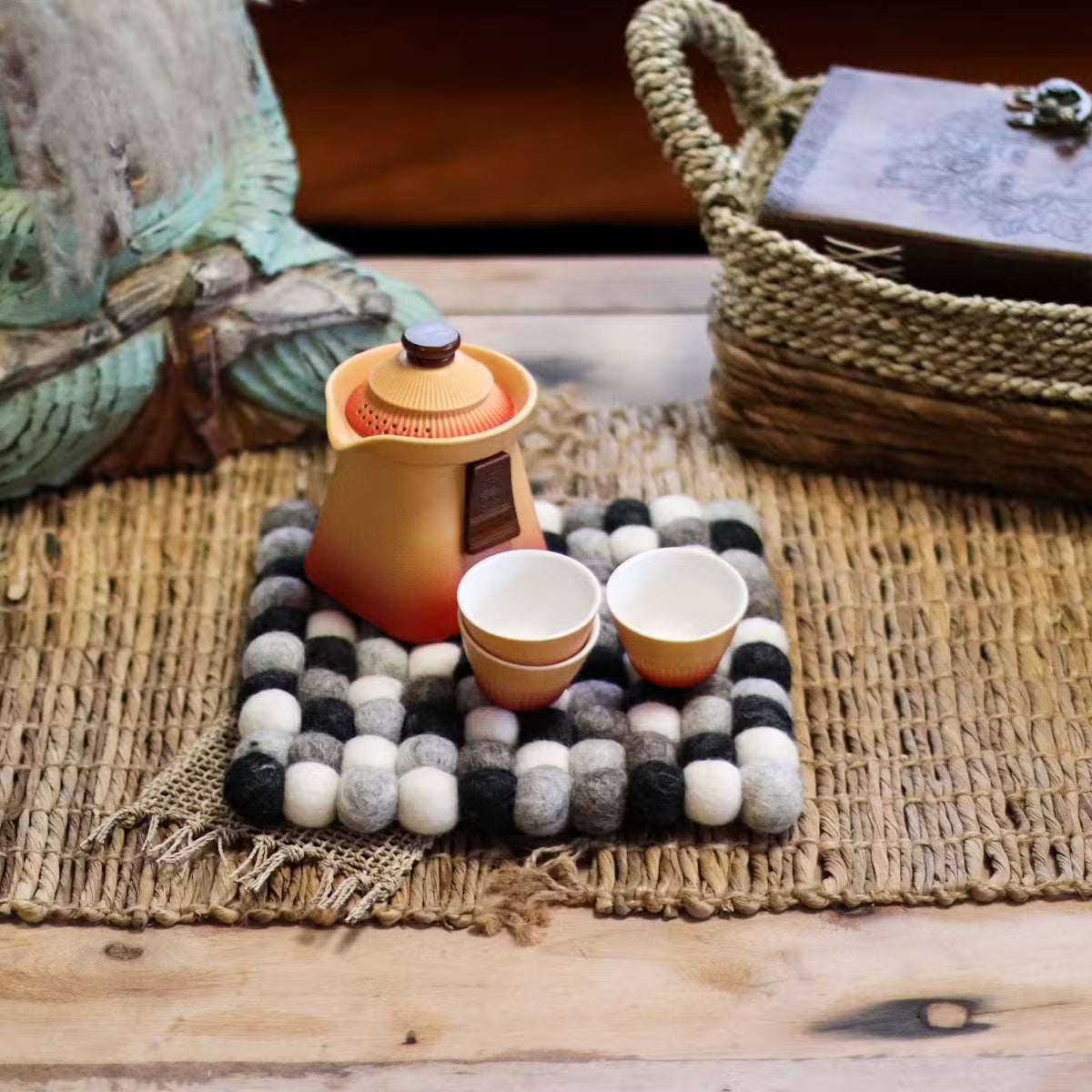 Grey felt ball square placemat under an orange teapot and two cups on a woven natural mat