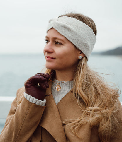 Woman wearing a beige headband, brown coat, and gloves by a body of water.