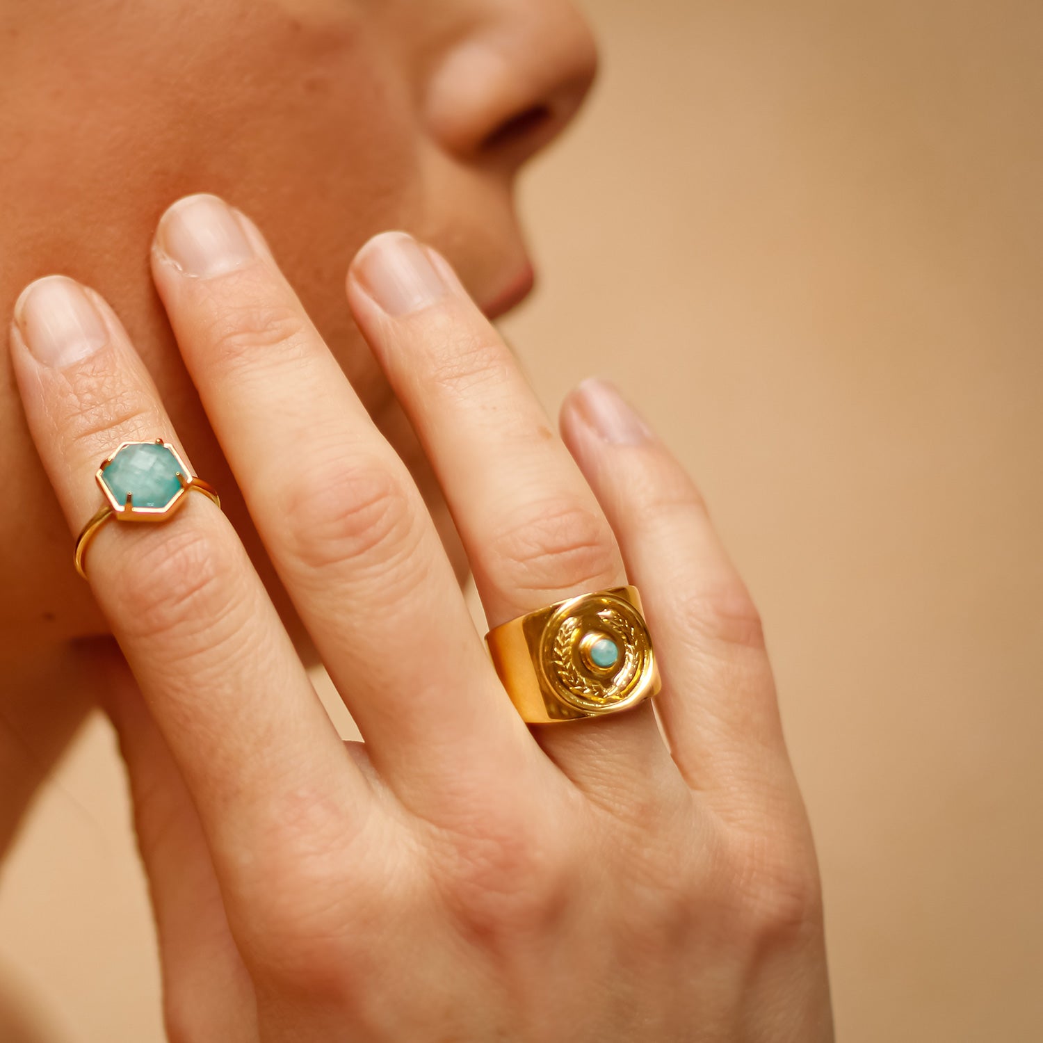 Close-up of a hand wearing two gold rings with turquoise stones on a beige background