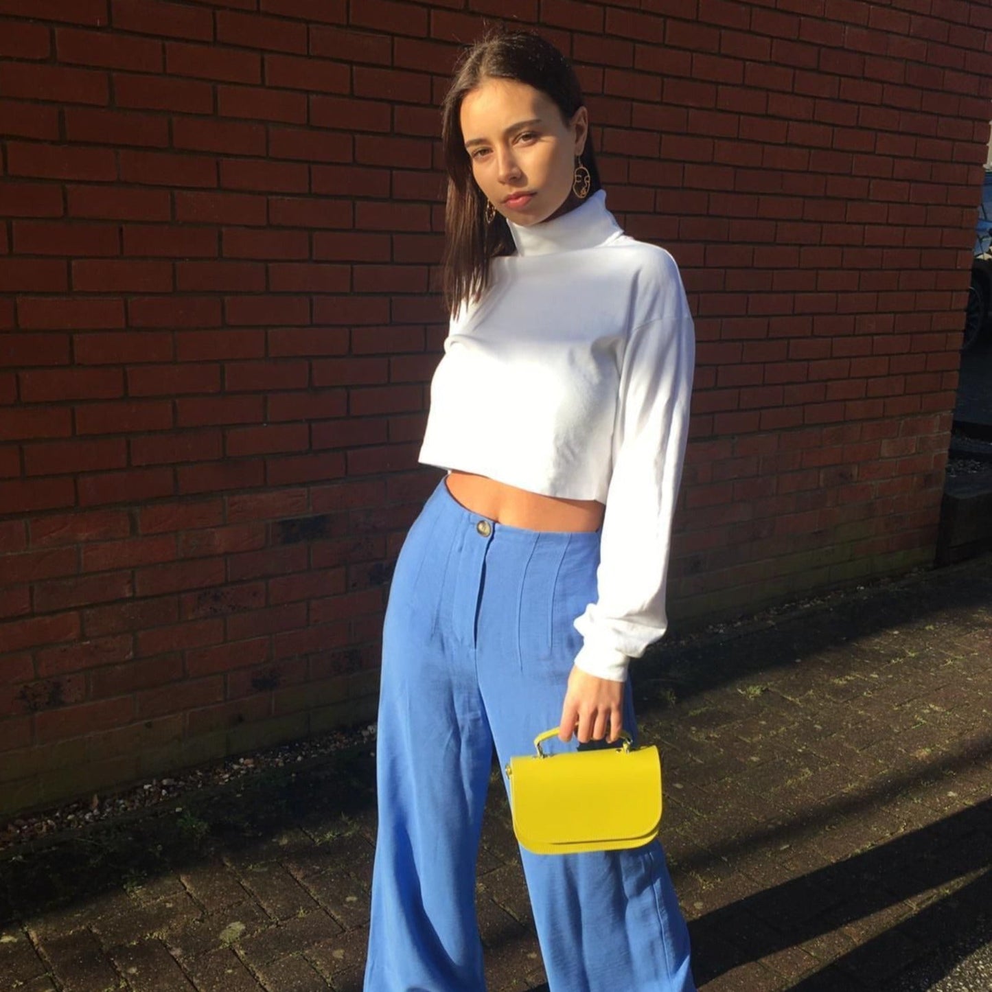Woman in white cropped top and blue pants holding a yellow handbag against a brick wall.